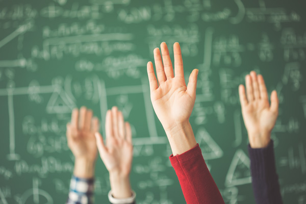 4 Hände melden sich in einem Klassenzimmer vor einer beschriebenen grünen Tafel
